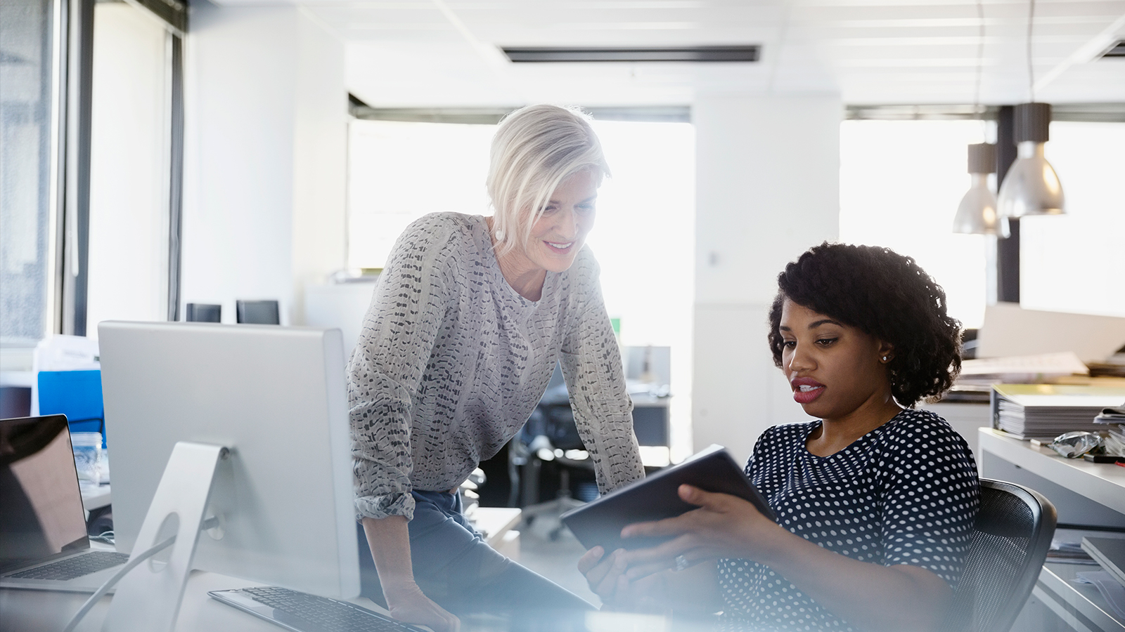 Woman sitting at a desk showing a tablet to another woman who is standing next to her.